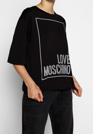 Persoon draagt een losse zwarte korte mouw T-shirt met witte omtrek en de tekst "LOVE MOSCHINO", gecombineerd met een donkere jeans.