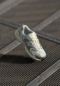 Witte en grijze New Balance hardloopschoen met veters, rustend op een gestructureerd metalen roosteroppervlak.