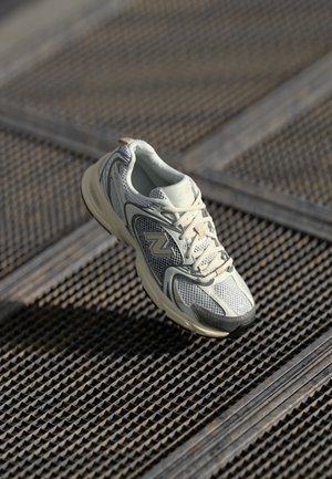 Witte en grijze New Balance hardloopschoen met veters, rustend op een gestructureerd metalen roosteroppervlak.
