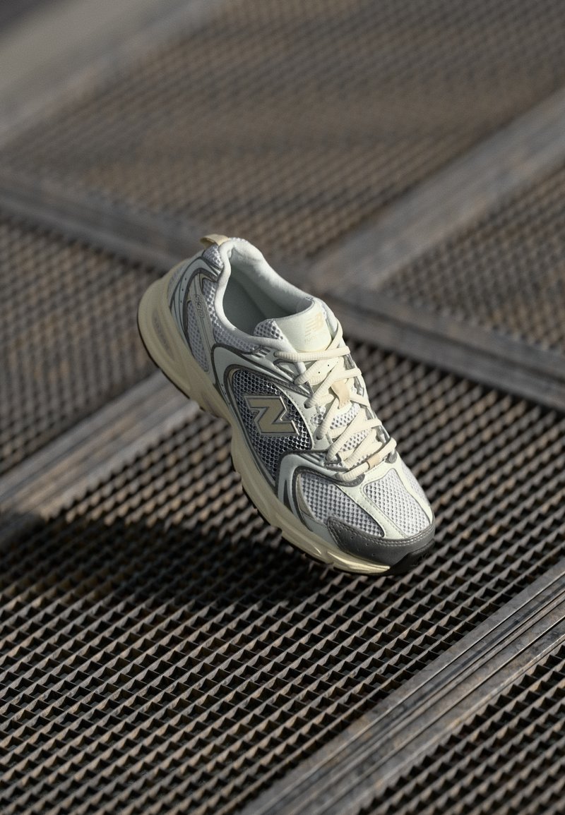 Witte en grijze New Balance hardloopschoen met veters, rustend op een gestructureerd metalen roosteroppervlak.