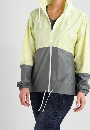Personne portant une veste Columbia zippée bicolore jaune et grise avec cordons blancs et poignets élastiques, associée à un jean bleu foncé.