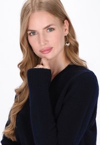 Maglione blu navy con polsini a coste, caratterizzato da un design a V. Modello con orecchini pendenti con perla, capelli lunghi e ondulati.