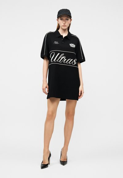 Γυναίκα που φοράει μια μαύρη υπερμεγέθη πόλο φόρεμα με το κείμενο "Ultras", μαύρο καπέλο και μαύρες ψηλοτάκουνες γόβες, στέκεται μπροστά σε λευκό φόντο.