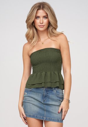 Jeune femme aux longs cheveux blonds portant un haut vert sans bretelles et une jupe en denim bleu, debout devant un fond clair uni.