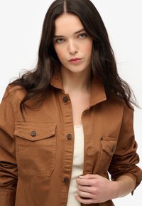 Brązowa koszula zapinana na guziki wykonana z bawełny, z dwiema kieszeniami na piersi i klasycznym kołnierzykiem, noszona na jasnym topie na ramiączkach. Gładka tekstura.