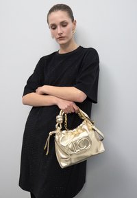 Bolso metálico dorado con un diseño suave y fruncido, acento de cadena y detalle de logotipo. La modelo lleva un vestido negro texturizado.