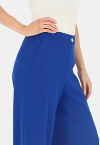 Niebieskie spodnie szerokie wykonane z gładkiego materiału. Posiadają płaski pas z małym złotym ozdobnym akcentem oraz kieszenie boczne.