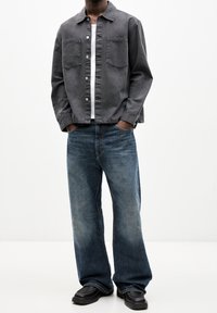 Hombre vestido con chaqueta abotonada gris oscuro, camiseta blanca debajo, jeans azules holgados y zapatos negros, de pie con las manos en los bolsillos.