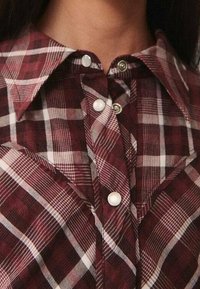 Košile s burgundským kostkovaným vzorem, klasickým límcem, bílými knoflíky a měkkou, texturovanou tkaninou. Výrazné diagonální a horizontální pruhy v designu.