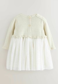 Langemouwige jurk met een gebreide beige top, hartvormige knopen op de rug en een witte tule rok met decoratieve stippen en ruffles.