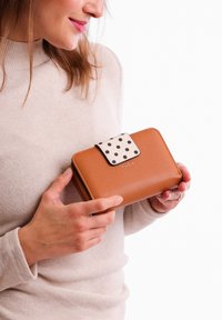 Brown leather wallet with a contrasting white flap featuring black polka dots, zip closure, and subtle branding embossed on the front.