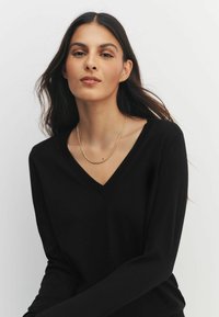 Schwarzer V-Ausschnitt-Pullover aus weichem Material mit langen Ärmeln und glatter Textur. Eine goldene, verdrehte Kettenhalskette schmückt den Ausschnitt.