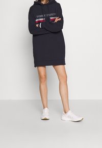 Mörkblå hoodieklänning med broderad logotyp, randiga detaljer i rött och vitt, ribbade muddar och sidoknappar, tillsammans med vita sneakers.