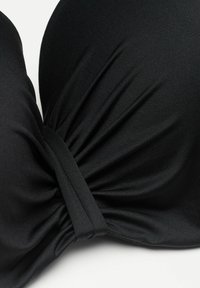 Gros plan sur un tissu noir torsadé au centre, montrant une texture côtelée et un détail froncé sur un fond uni.