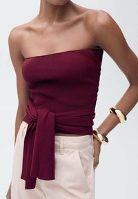 Bordeaux geribbelde strapless top met een strik in de taille, gecombineerd met lichtroze broeken en gouden ronde armbanden. Glad materiaal, eenvoudig ontwerp.