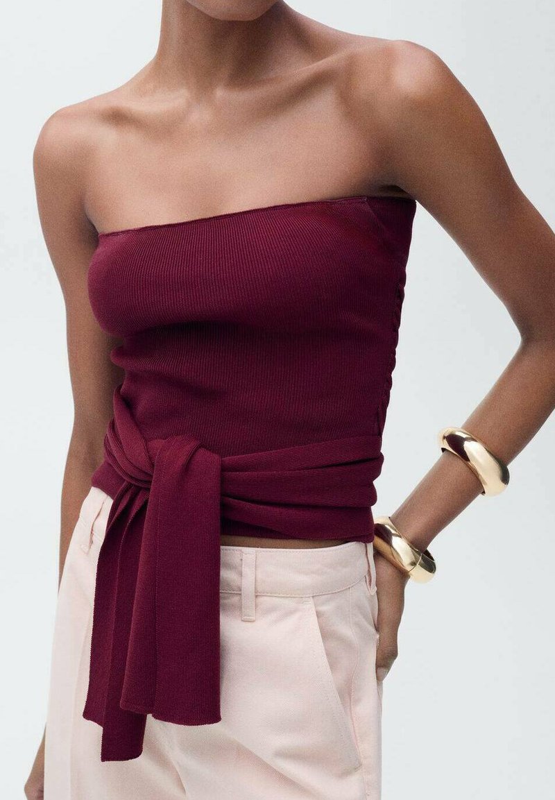 Bordeaux geribbelde strapless top met een strik in de taille, gecombineerd met lichtroze broeken en gouden ronde armbanden. Glad materiaal, eenvoudig ontwerp.