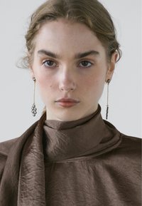 Boucles d'oreilles longues de couleur dorée ornées de grappes de petites pierres précieuses sombres, portées avec un haut marron texturé à col montant.