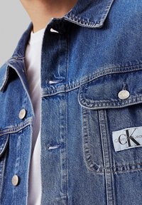 Džínová bunda ve světle modré barvě se stříbrnými cvoky, dvěma náprsními kapsami a štítkem s logem CK. Má klasický límec a detaily v prošívání.