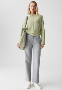 Vrouw in een lichtgroene trui, grijze rechttoe-rechtaan jeans, witte sneakers en met een gevoerde lichtgroene schoudertas.