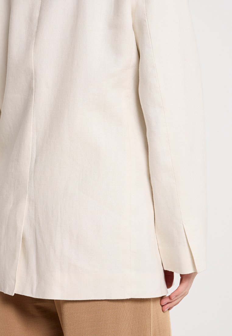 Blazer beige clair en lin, avec une coupe droite, une fente au dos et des détails de couture visibles. Texture lisse et design minimaliste.