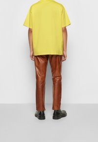 Camiseta amarilla de gran tamaño y mangas cortas, combinada con pantalones de cuero ajustados marrones y botas negras al tobillo, vista desde atrás sobre un fondo neutro.