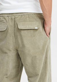 Khakifarbene Cordhosen mit vertikalen Rillen, elastischem Bund, zwei Gesäßtaschen und einem kleinen Etikett an der rechten Tasche.