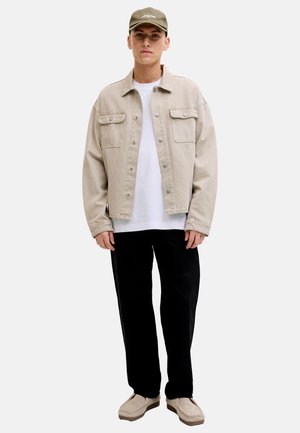 Young man wearing beige cap, beige button-up jacket, white t-shirt, black pants, and beige shoes, standing against plain white background.