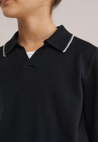 Femme portant un polo en maille noire avec des surpiqûres blanches le long du col, profil visible du cou et d'une partie du visage.