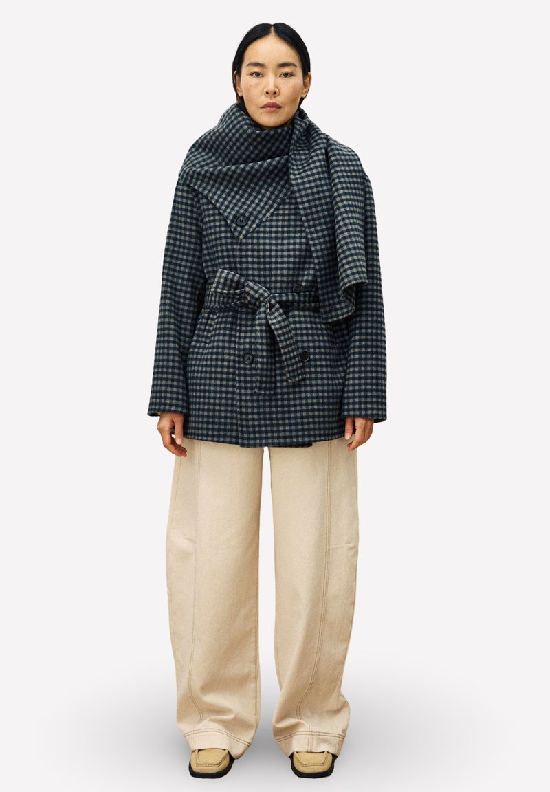 Manteau en mélange de laine, à carreaux bleu et gris avec un large col et une ceinture, associé à un pantalon large crème et des baskets beiges.