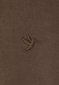 Der braune Stoff weist ein strukturiertes, geripptes Muster auf, verziert mit einem kleinen, gestickten Kolibri-Logo in einem etwas dunkleren Braunton.