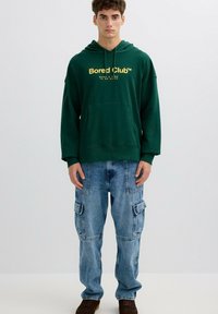 Sudadera con capucha verde con texto dorado, combinada con jeans cargo azul claro con múltiples bolsillos y un ajuste relajado. Zapatos marrones completan el conjunto.