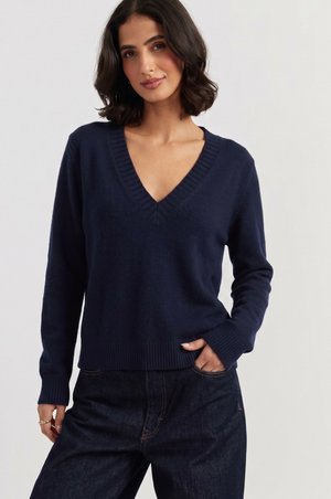Vrouw draagt een marineblauwe V-hals trui en donkere spijkerbroek, met één hand in haar zak, tegen een effen lichte achtergrond.