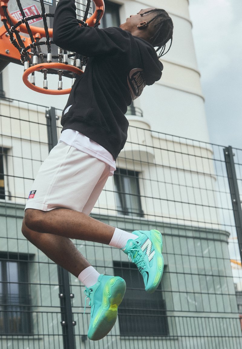 Sportbekleidung mit einem schwarzen Kapuzenpullover mit Logo, weißen Mesh-Shorts und leuchtend türkisfarbenen Sneakers mit schwarzen und gelben Akzenten, vor einem Basketballplatz-Hintergrund.