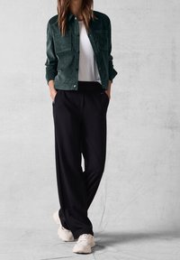 Dark green corduroy jacket with two chest pockets, paired with black wide-leg trousers and light sneakers, against a neutral background.