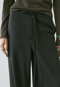 Kvinde iført sorte høj-taljede bukser med forreste knapper og snøre, brun langærmet top, armbånd på højre håndled og ringe på venstre hånd.