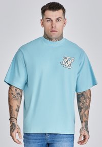 Világoskék, oversized póló, amely pamutból készült, kerek nyakkivágással és fehér, stilizált logóval a bal mellrészen.