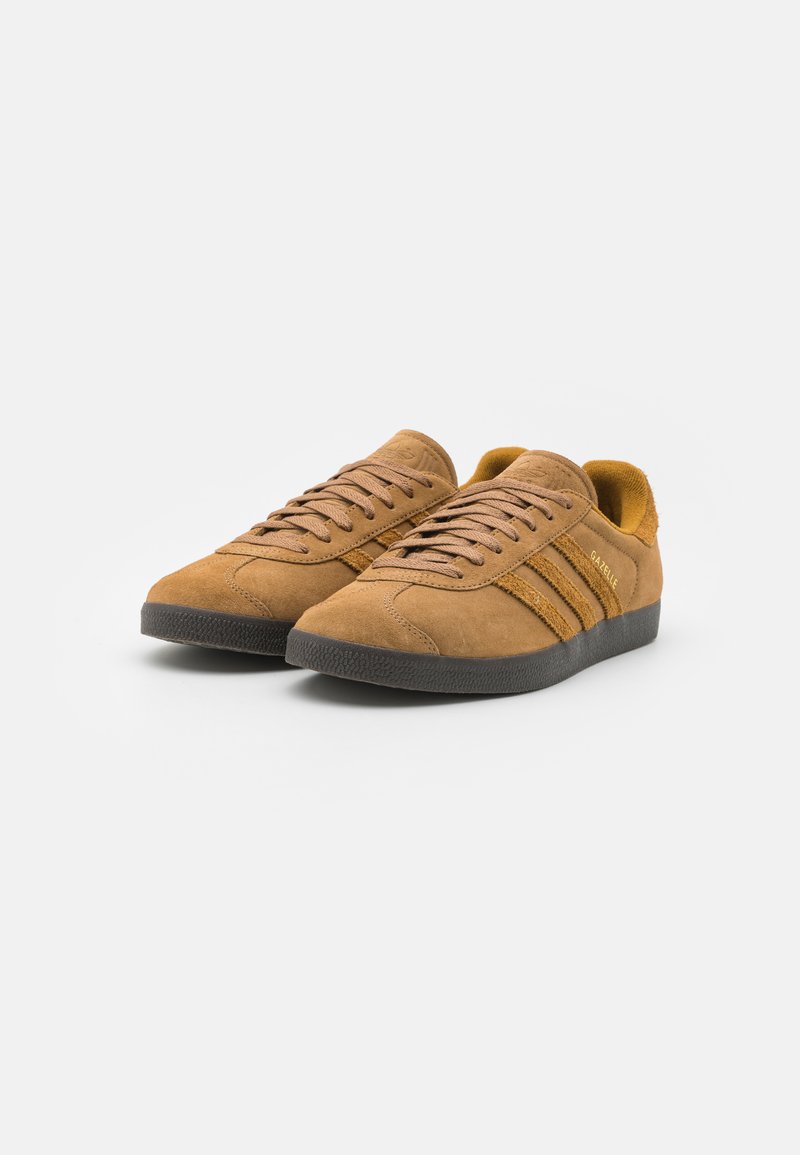 Zamszowe sneakersy w jasnobrązowym kolorze z tonalnym systemem sznurowania, z dwoma ciemnobrązowymi paskami po bokach i czarną gumową podeszwą.