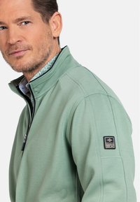 lichtgroene quarter-zip jas met een gestructureerde afwerking, voorzien van een zwarte ritssluiting en een logo patch op de linkermouw. Met kraag ontwerp.