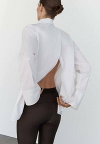 Camisa blanca con la espalda descubierta y mangas largas, combinada con leggings negros. La tela parece ligera y con una textura suave.