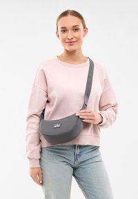 Szara tkaninowa torba na ramię o zaokrąglonym kształcie i regulowanym pasku, z czarną metką i gładką fakturą, noszona przez modelkę w jasnoróżowej bluzie.