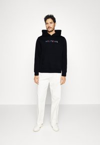 Black hoodie made of cotton with a front pocket and embroidered multi-colored "HILFIGER" logo. Paired with white trousers and sneakers.