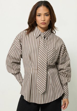 Jeune femme aux cheveux bruns porte une chemise oversize rayée marron et blanc avec une cravate assortie, debout sur un fond uni.