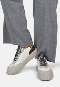 Sneakers in pelle bianchi, grigi e bronzo con dettagli testurizzati, una suola spessa e lacci. Pantaloni a gamba larga grigi parzialmente visibili.