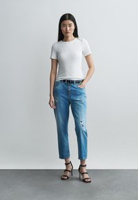 Jauna moteris stovi priešais paprastą foną, vilkėdama baltą trumpomis rankovėmis marškinėlį, suplėšytus mėlynus džinsus, juodą diržą ir juodus aukštakulnius sandalus.