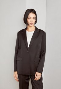Black tailored blazer made of smooth fabric, featuring a single button, notch lapels, and two front pockets. Paired with a white top.