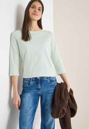 Frau mit einem mintgrünen und weiß gestreiften Oberteil und blauer Jeans, die eine braune Jacke mit Struktur hält, steht vor einer weiß-beigen Wand.