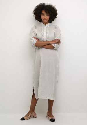 Robe longue rayée en crème et gris clair, avec un col à volants, des manches trois-quarts et des fentes sur les côtés. Complétée par des talons beiges avec des bouts noirs.