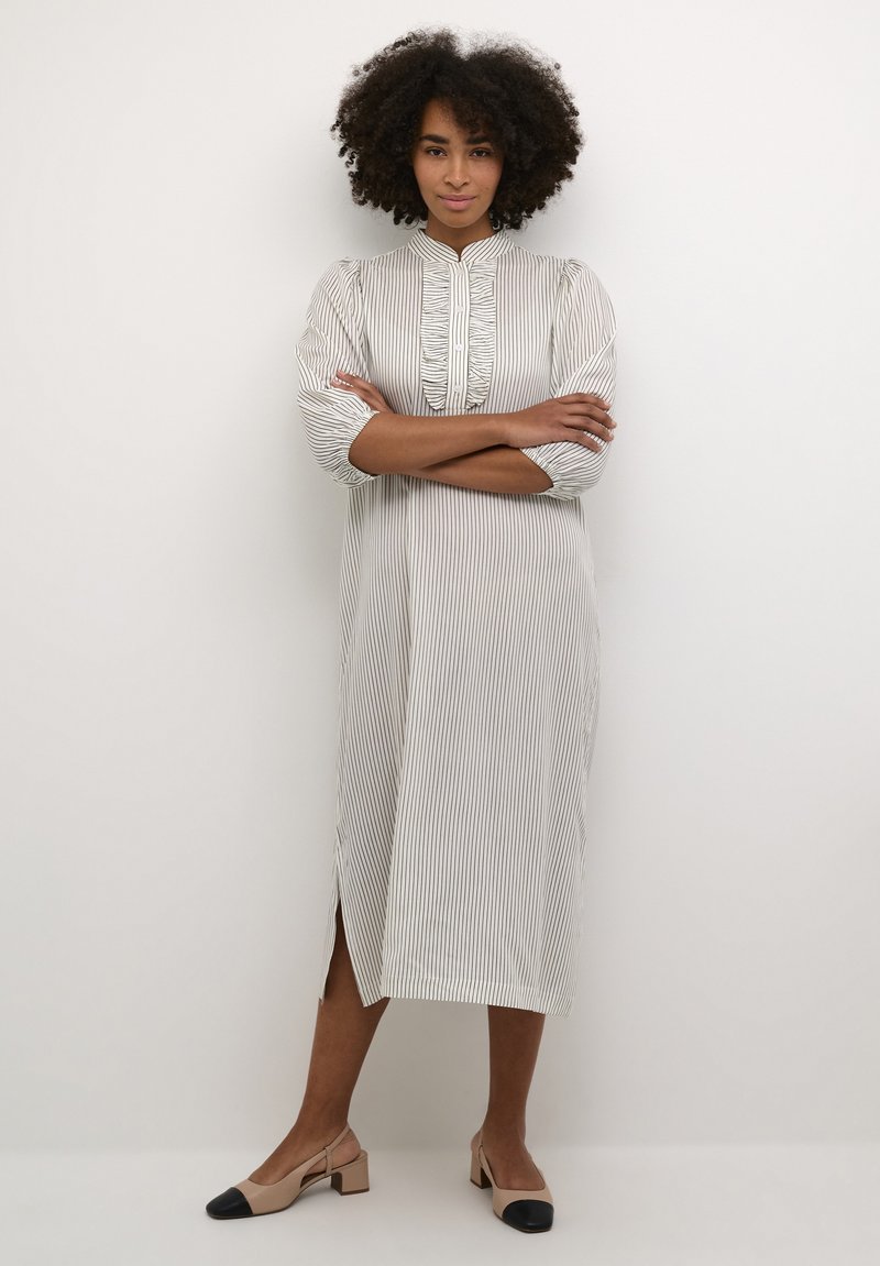 Robe longue rayée en crème et gris clair, avec un col à volants, des manches trois-quarts et des fentes sur les côtés. Complétée par des talons beiges avec des bouts noirs.