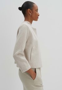 Helle beige, kurz geschnittene Jacke aus weichem Material mit rundem Ausschnitt und Seitentaschen, kombiniert mit hellen, taillierten Hosen.