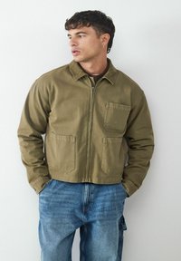 Olive green, zip-front jacket made of sturdy fabric, featuring two front pockets and a classic collar. Paired with blue denim jeans.
